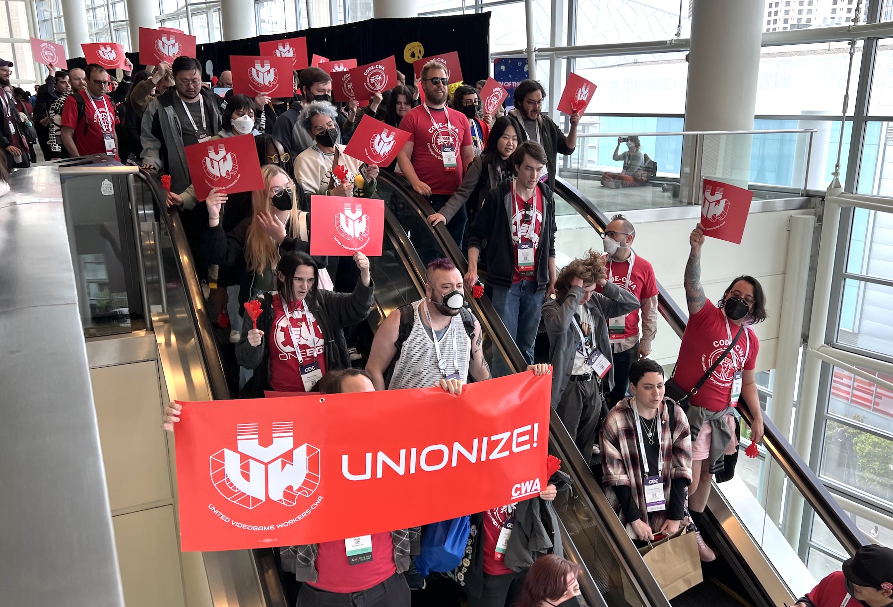 Game workers march down the escalator at GDC Workers wearing GDC conference passes fill an escalator going down. Many hold signs that say UVW and one holds a larger banner that says UVW: UNIONIZE!