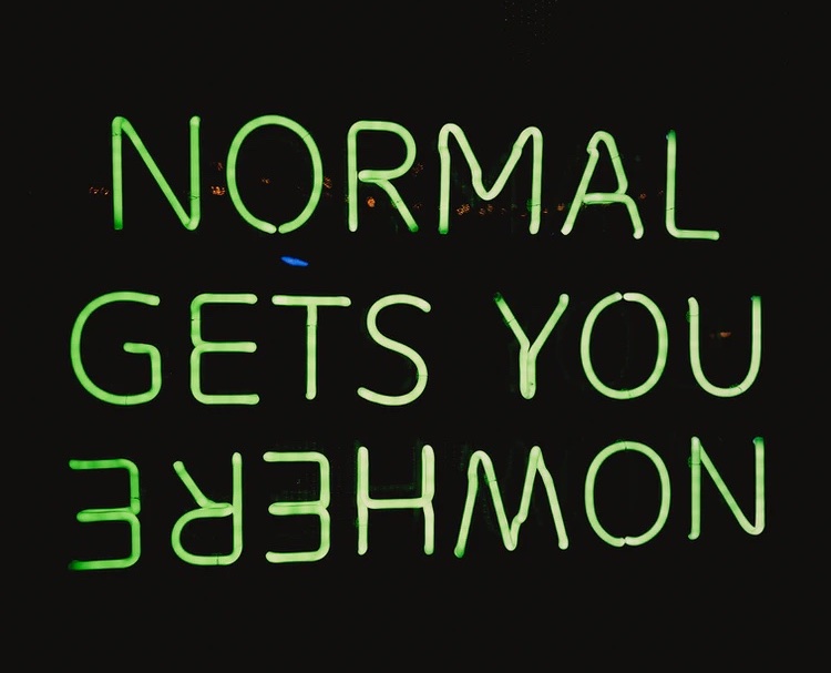 A neon green sign on a black background that says NORMAL GETS YOU NOWHERE, with NOWHERE upside down and backwards