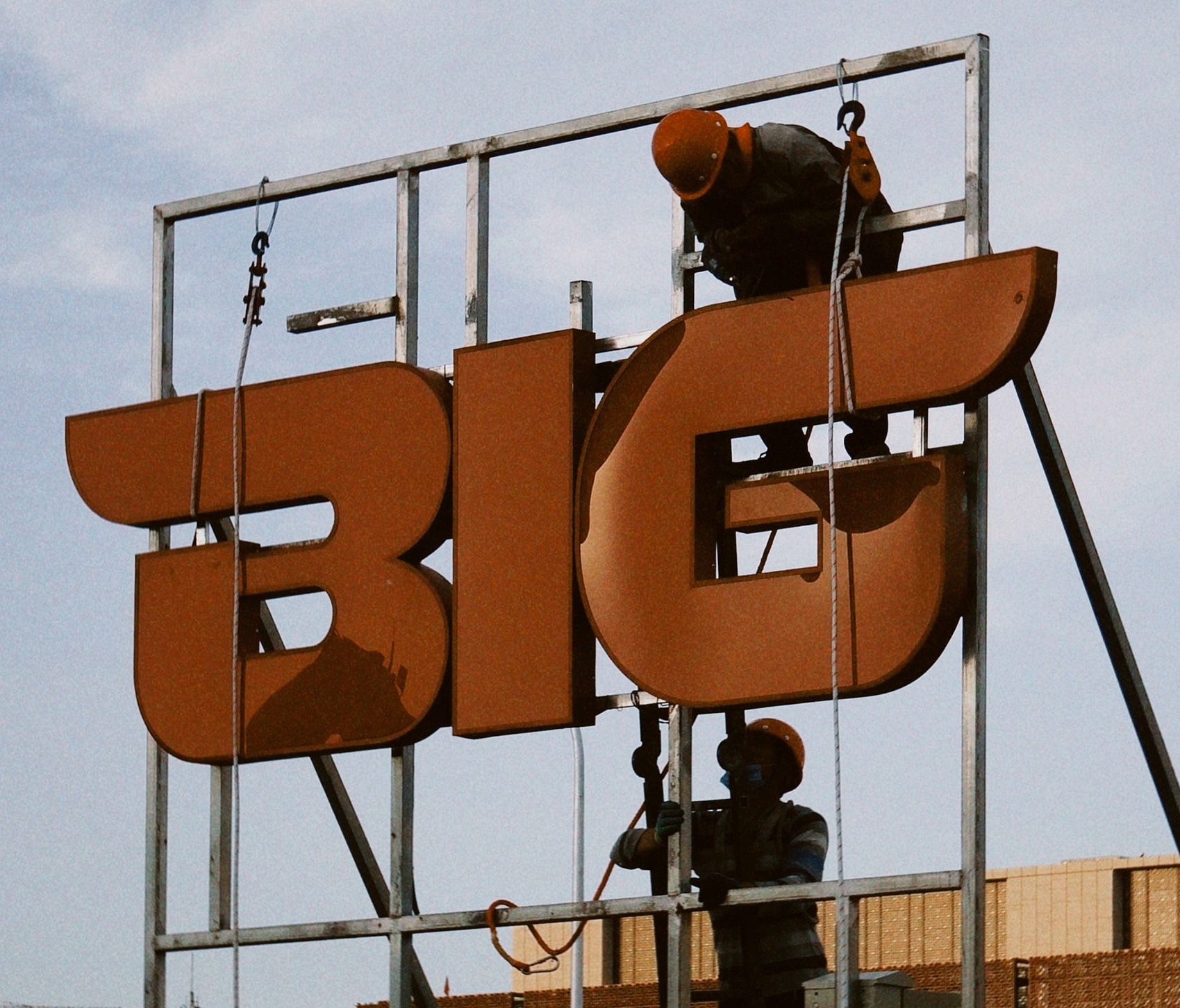 Two workers on the job, talking it out as they install an organgle rooftop sign that says BIG